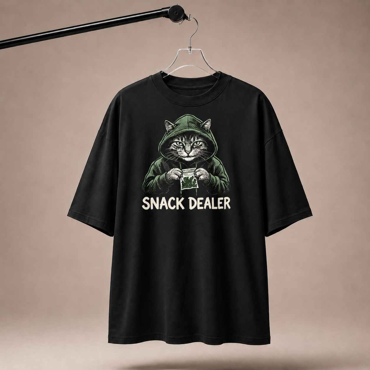 Black t-shirt with a graphic of a cat holding a snack and 'SNACK DEALER' text, hanging on a beige background.
