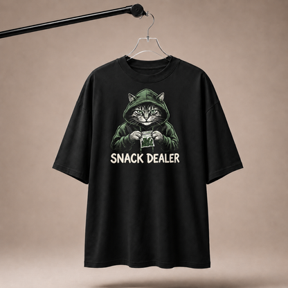 Black t-shirt with a graphic of a cat holding a snack and 'SNACK DEALER' text, hanging on a beige background.
