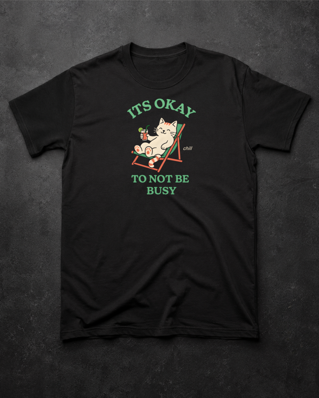 A black short-sleeve graphic t-shirt laid flat on a dark textured background. The shirt features a cartoon illustration of a white cat lounging in a red beach chair, with green text reading 'ITS OKAY TO NOT BE BUSY'.